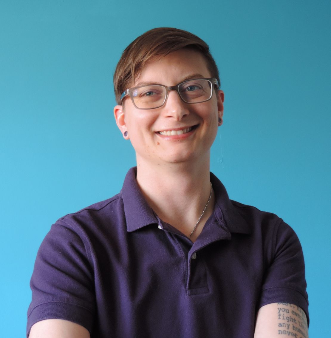 Massage therapist with brown hair wearing a purple polo shirt, gray glasses with part of a tattoo showing on their arm. They are against a blue background and smiling at the camera.